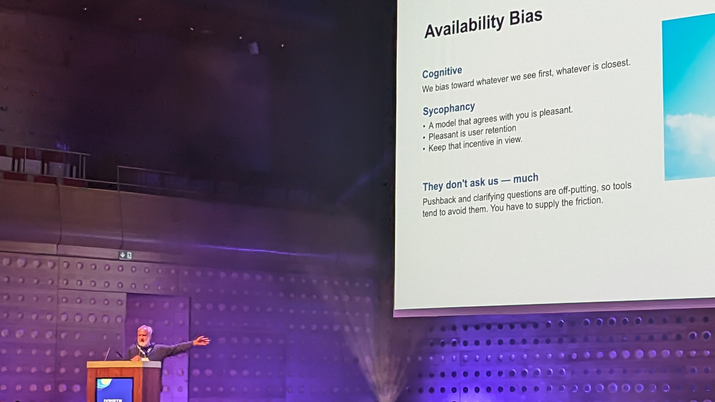 Michael at the bottom left, the slide behind him saying:
Availability Bias
Cognitive
We bias towards what we see first, whatever is closest
Sycophancy
- a model that agrees with you is pleasant.
- pleasant is user retention.
- keep that incentive in view.
They don't ask us - much
Pushback and clarifying questions are off-putting, so tools tend to avoid them. You have to supply the friction